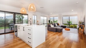 A modern kitchen with a large island, a dining area with a table and chairs, a gray sofa, and large windows with a view of greenery. - Pet-friendly. Bright and Spacious Dunsborough Home (Dunsborough)