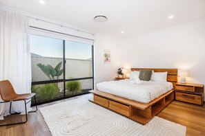 Quarto com uma grande janela, uma cama com cabeceira de madeira, mesas de cabeceira e uma cadeira.