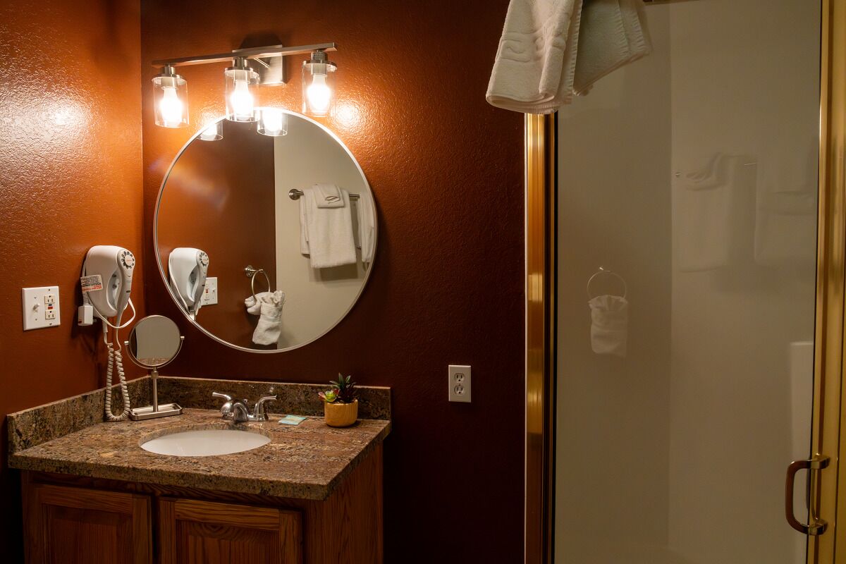 A bathroom with a granite countertop sink, a round mirror, a towel rack, a handheld mirror, a hairdryer, a hair straightener, and a potted plant.