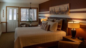 A bedroom with a large bed, a nightstand with a lamp, a chair, a window with blinds, and a door.