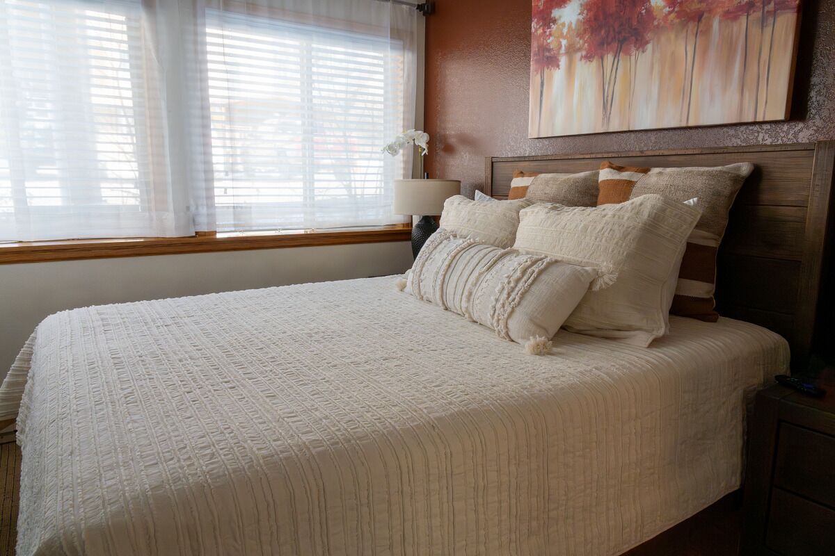 A neatly made bed with a white quilt and pillows, a bedside lamp, and a painting on the wall.