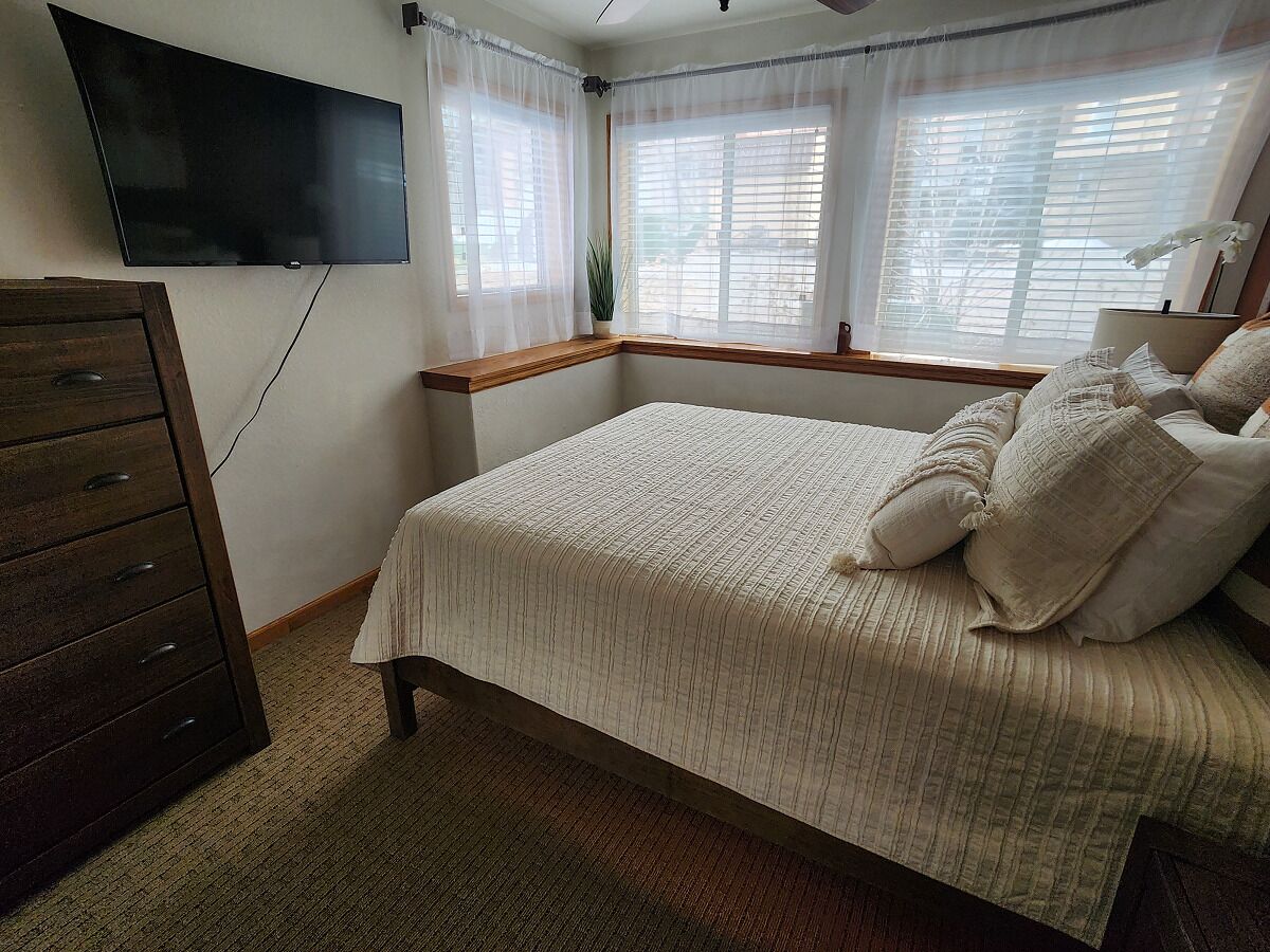 Une chambre à coucher comprenant un lit, une commode, un téléviseur fixé au mur et des fenêtres équipées de stores.