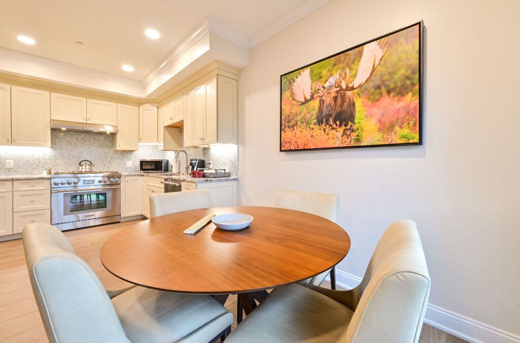 A modern kitchen with white cabinets, a stainless steel oven, and a microwave. There is a round wooden table with two chairs in the foreground.