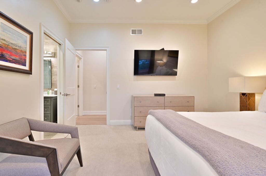 A bedroom with a bed, a dresser, a chair, and a TV mounted on the wall.