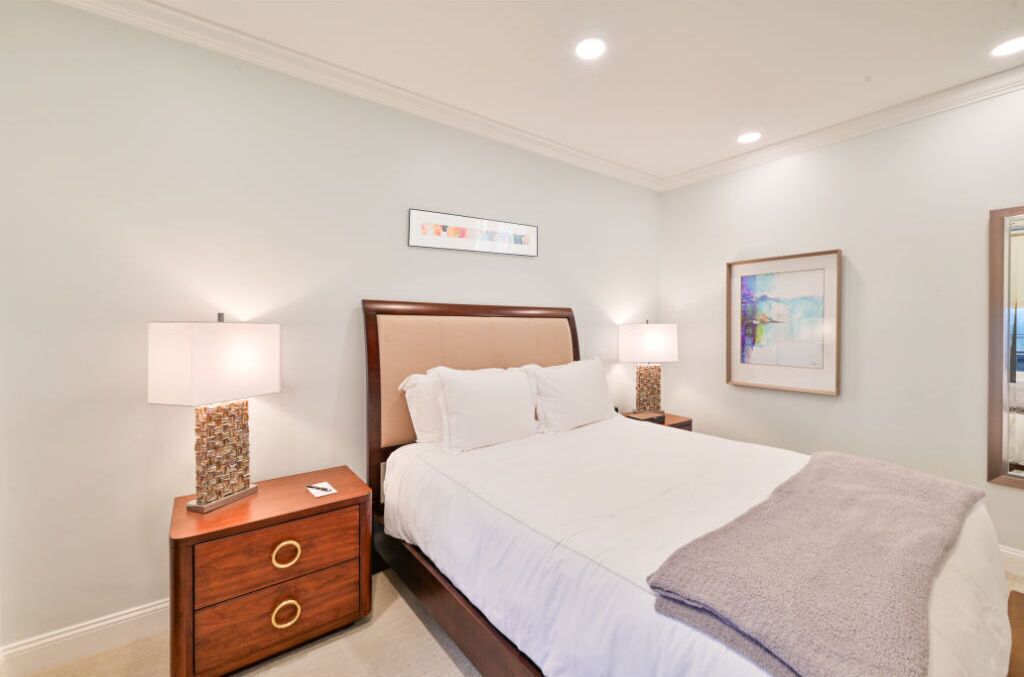 A neatly arranged bedroom with a bed, two bedside tables with lamps, a small wooden dresser, and a framed picture on the wall.