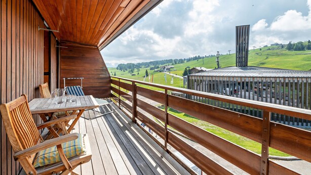 Una terraza de madera con una mesa y sillas, con vista a un paisaje verde y un edificio a lo lejos.
