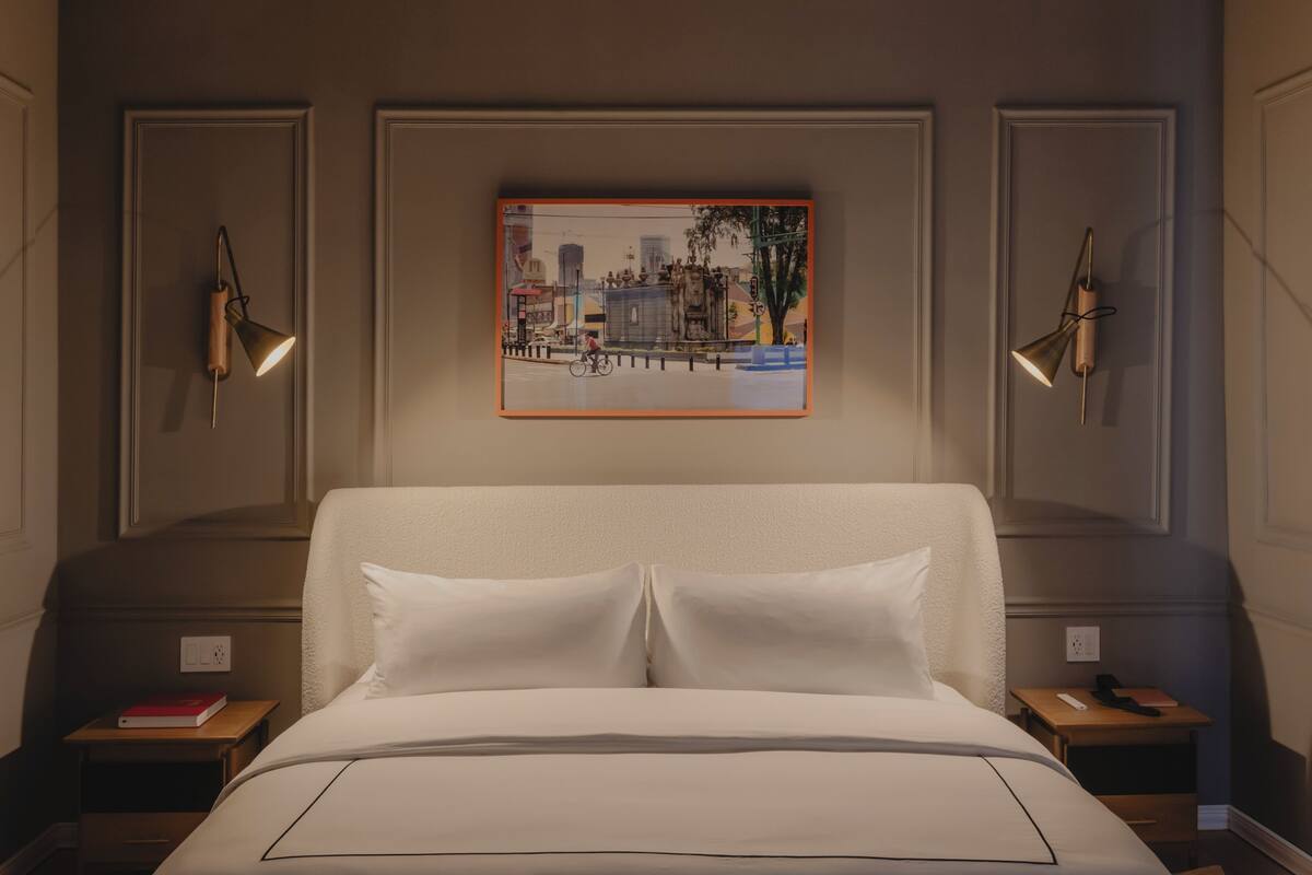 A hotel room with a large bed, two bedside tables, a framed cityscape artwork, and two wall-mounted lamps.