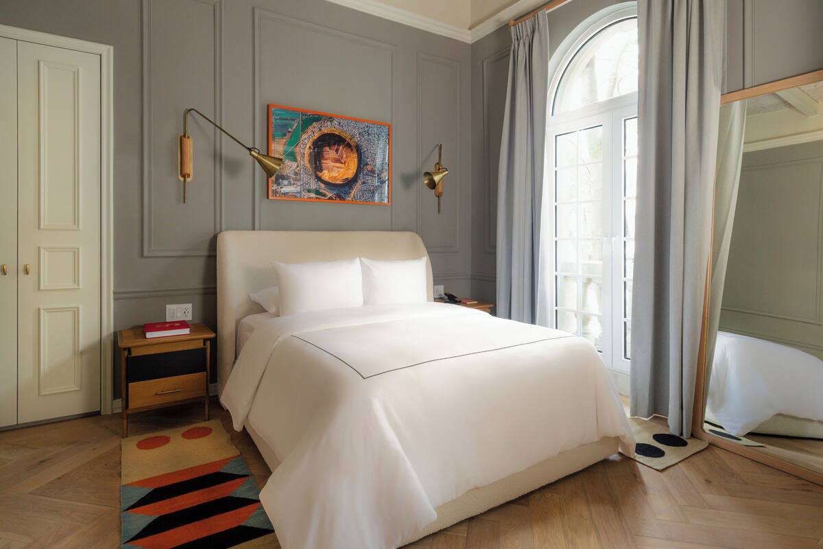 A neatly arranged bedroom with a large bed, a bedside table, a nightstand, a mirror, and a window with curtains.
