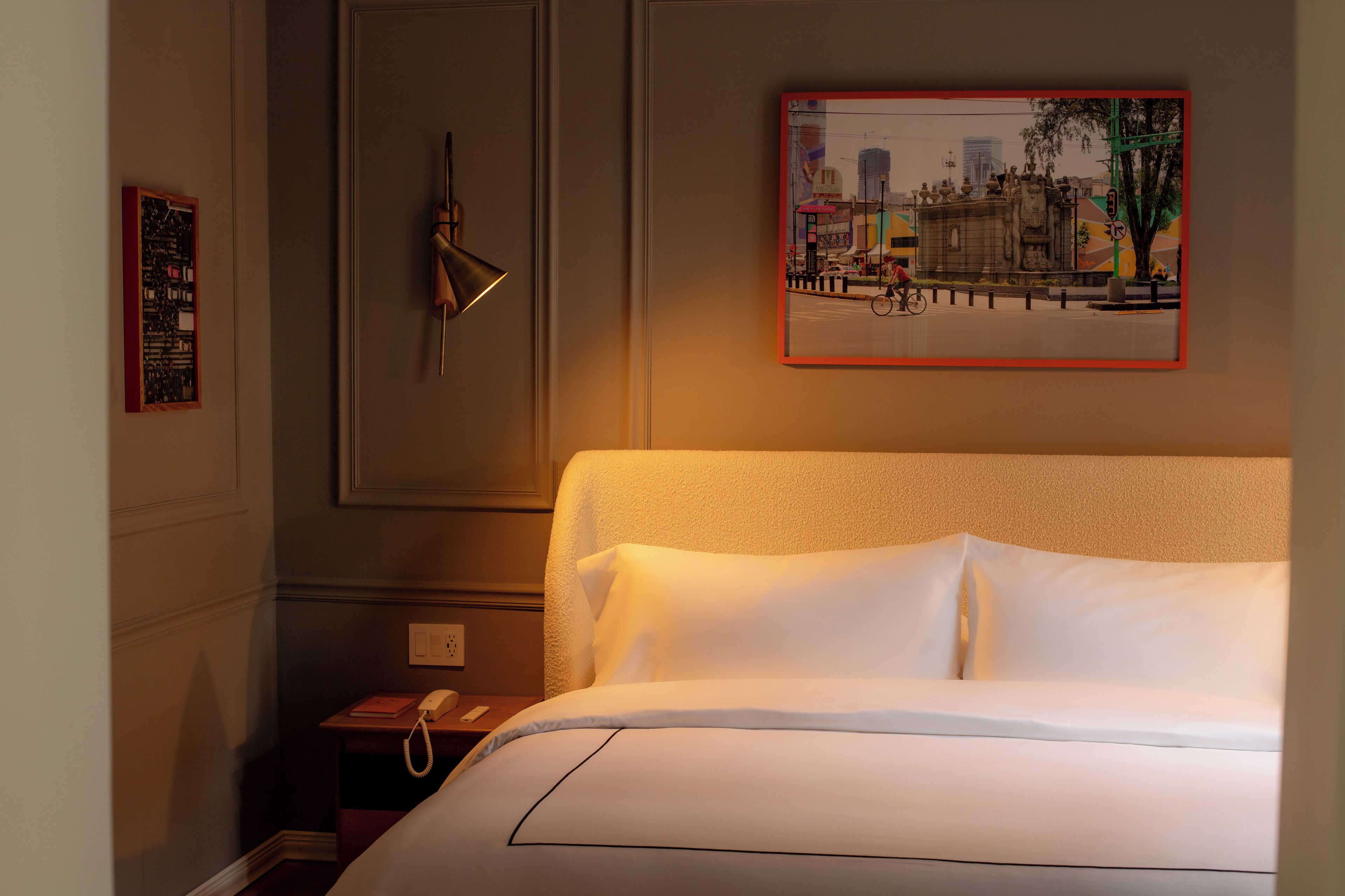 Une chambre d’hôtel avec un lit, une table de chevet avec un téléphone, un tableau encadré au mur et une porte avec une poignée.