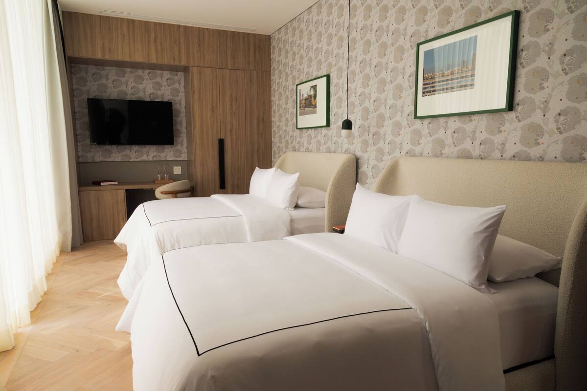 A hotel room with two beds, a flat-screen TV mounted on the wall, a small table with a lamp, and a framed picture on the wall.