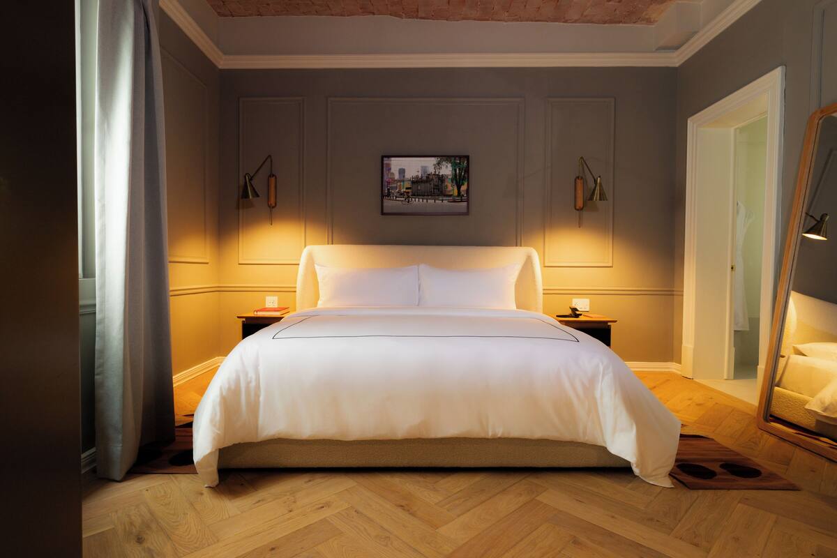 A neatly made bed in a hotel room with a cityscape painting on the wall, wooden flooring, and a mirror on the right.