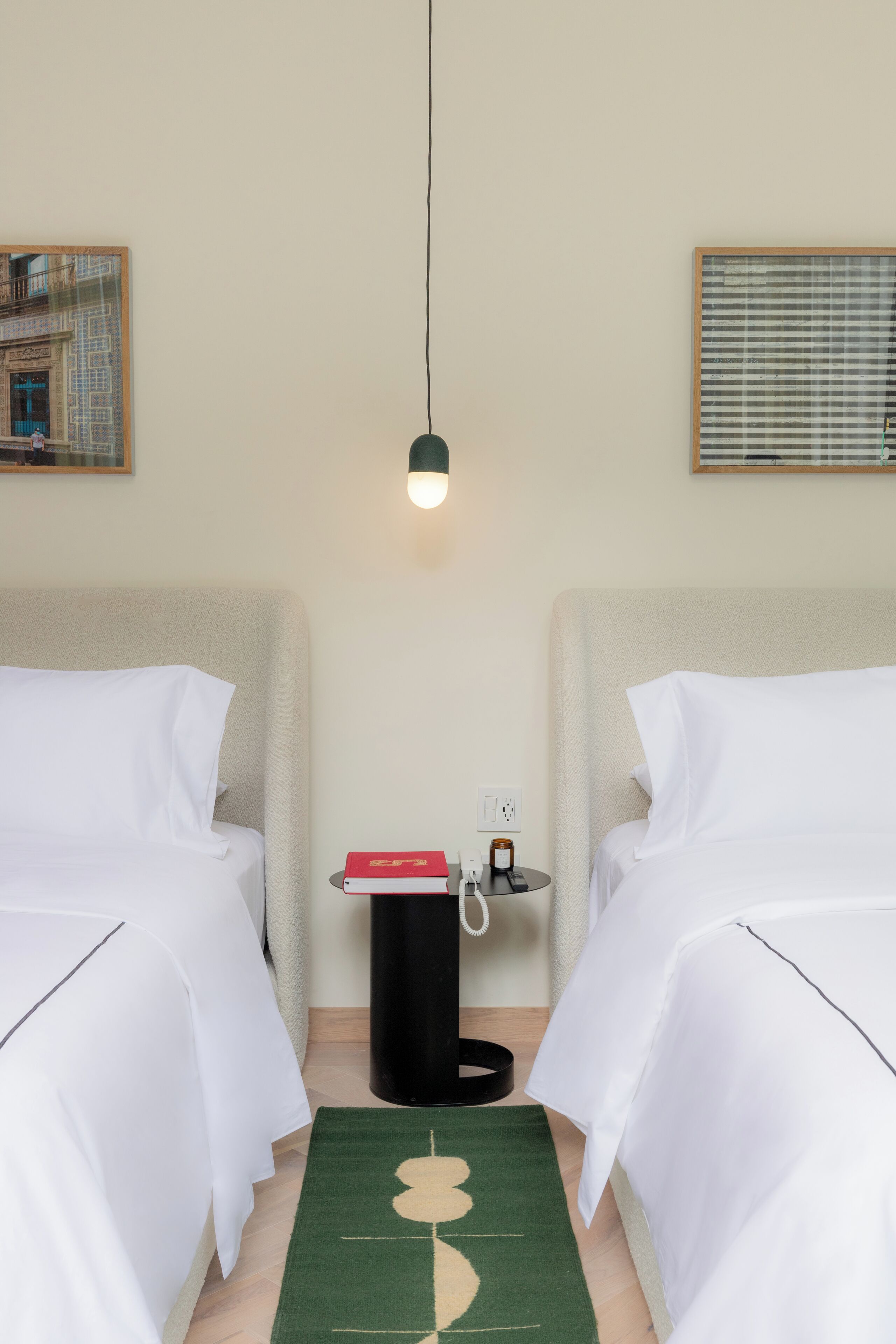 A hotel room with two beds, a green rug, a nightstand with a book and a lamp, and a wall-mounted light fixture.