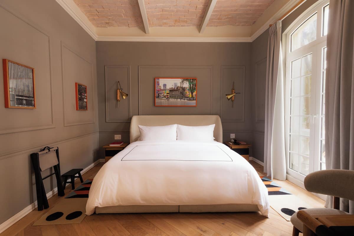 A neatly arranged bedroom with a large bed, a small table, and a chair. There is a window with curtains, a painting on the wall, and a wooden floor.