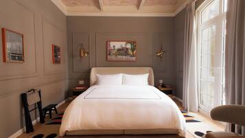 Una camera da letto ordinata con un letto grande, un tavolino e una sedia. Si vede una finestra con tende, un quadro appeso al muro e un pavimento in legno.