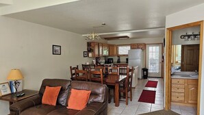 A living room with a brown leather sofa, wooden dining table, and chairs. A kitchen is visible in the background with wooden cabinets and a refrigerator. - Cascade Three Bedroom Two Bath C07 (Ouray)