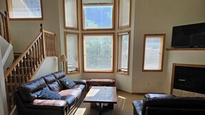 A living room with a leather sofa, a coffee table, and a TV. There are multiple windows with blinds, a staircase, and a fireplace. - Cascade Three Bedroom Two Bath C07 (Ouray)