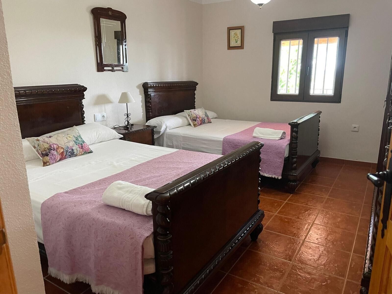 A hotel room with two wooden beds, a nightstand with a lamp, a mirror, a window, and a door.