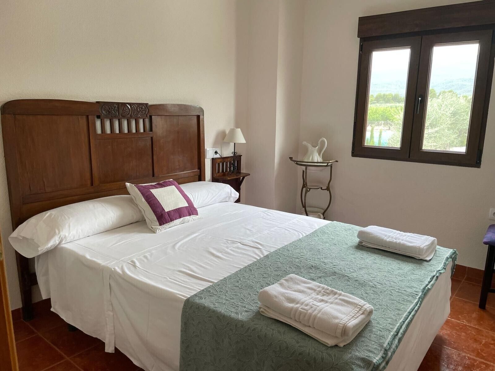 A neatly made bed with white linens and a green bedspread, a wooden headboard, a small bedside table with a lamp, and a window with a view of greenery.