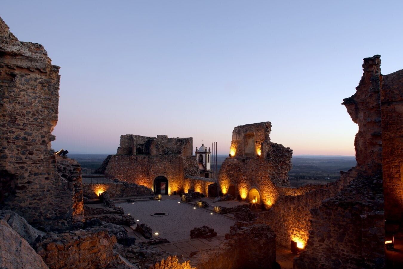 Una estructura de piedra antigua con ruinas, un patio central y una vista distante de un paisaje urbano.