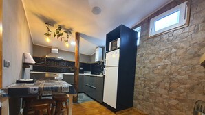 A compact kitchen with a wooden table, chairs, a refrigerator, and a stone wall.