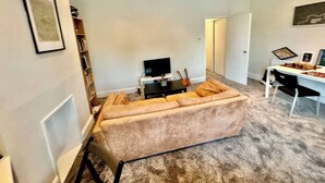 En stue med en beige sofa, et fjernsyn, en guitar og et spisebord med stole.