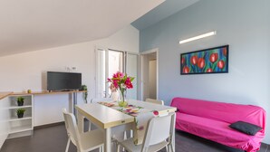 A modern living room with a dining table, chairs, a sofa, a flat-screen TV, a painting, and a vase with flowers.
