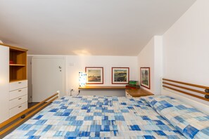 Ein Schlafzimmer mit einem Bett, einem Holzkopfstück, einem Nachttisch mit Büchern, einem Wandregal mit gerahmten Bildern und einer Tür.