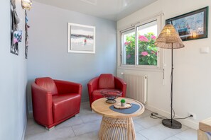 A room with two red leather chairs, a round wooden table with a small plant, a wicker floor lamp, a framed picture on the wall, and a window with flowers.