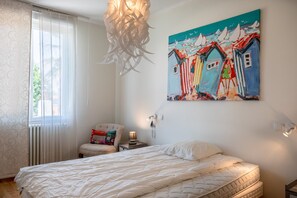 A bedroom with a bed, a chair, a painting, and a window with curtains.