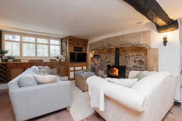 A cozy living room with a fireplace, a sofa, a television, and a kitchen area with wooden cabinets and a stone wall. - Cosy cottage on the outskirts of Bath (Saltford)