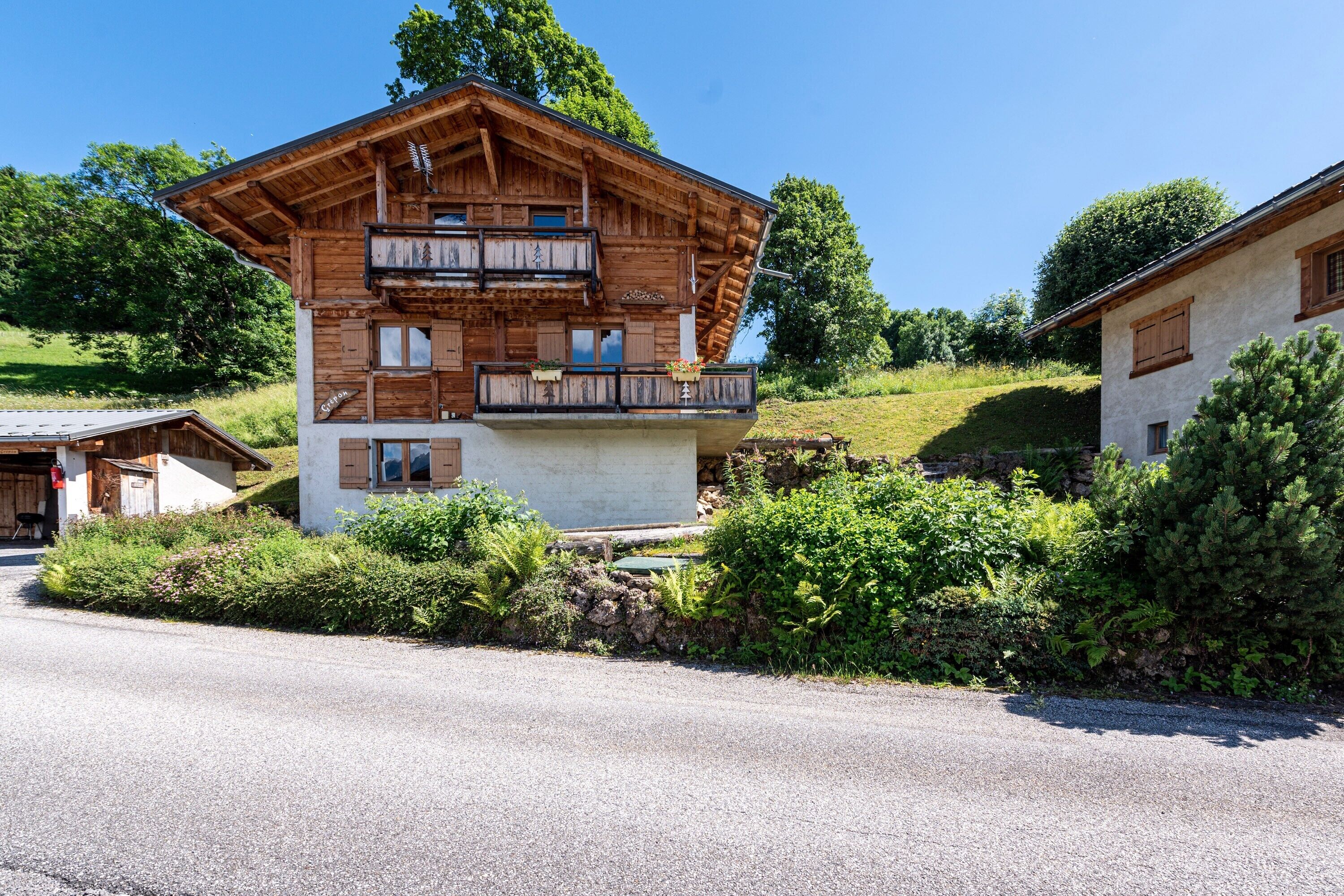 A two-story wooden chalet with a balcony, surrounded by greenery and a clear sky.
