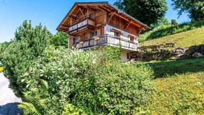 A wooden chalet with a balcony, surrounded by greenery and a stone wall.