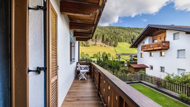 Een balkon met houten hekwerk, een tafeltje met een stoel, en uitzicht op een groen landschap met gebouwen en bomen.
