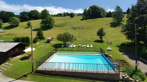 Una piscina rectangular con una valla alrededor, una pequeña construcción de madera y una colina cubierta de césped al fondo.