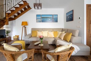 Una sala de estar con un sofá, dos sillones de mimbre, una mesa de centro, una lámpara, un televisor, una escalera y obras de arte en la pared.