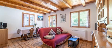 A cozy room with a wooden ceiling, a red sofa with pillows, a small table, and a window with a view of greenery.