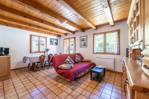 A cozy room with a wooden ceiling, a red sofa with pillows, a small table, and a window with a view of greenery.