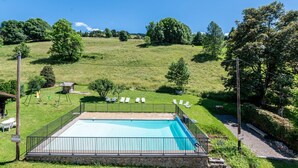 A swimming pool with a stone border, surrounded by a black fence, and a grassy area with trees and a hill in the background. - Apartment 'L'Hermine, Piscine Et Aire De Jeux' with Mountain View, Private Terrace and Wi-Fi (Crest-Voland)