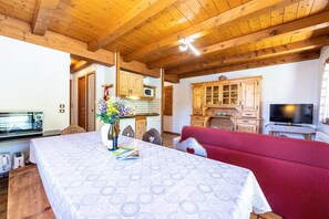 A room with a bed, a TV, wooden beams, a kitchen area, and a dining table.