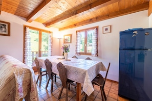 A dining area with a table set for six, wooden chairs, a refrigerator, and a window with curtains.