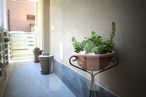 Un couloir avec une plante en pot posée sur un support métallique, un sol carrelé et un escalier en arrière-plan.