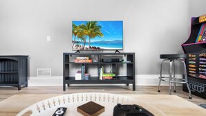 Ein Wohnzimmer mit Fernseher, der eine Strand Szene zeigt, einer Spielkonsole und einem Barhocker.