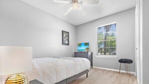 Ein Schlafzimmer mit einem Bett, einem Nachttisch, einer Lampe, einem Deckenventilator, einem Fenster mit Jalousien und einem kleinen Hocker.