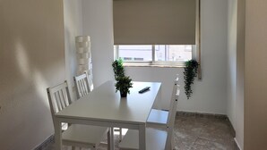 A corner room with a white table, chairs, a window with a blind, and a potted plant.