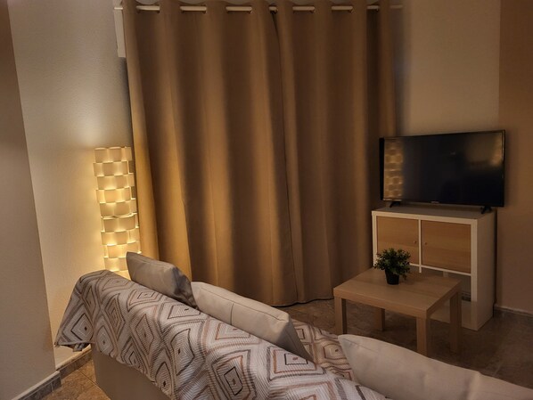 Un salon avec un canapé, un téléviseur posé sur un meuble, une lampe et un rideau.