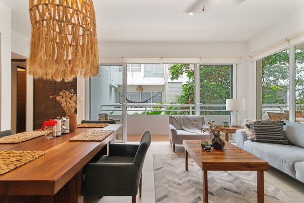 A modern living room with a wooden dining table, a sofa, and a coffee table. There is a large window with a view of greenery outside.