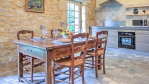 A rustic stone wall, a wooden dining table with chairs, a fruit bowl, wine glasses, and a kitchen with a microwave and oven. - Holiday Home 'Le Bel Prat' with Private Pool, Private Terrace and Wi-Fi (Domme)