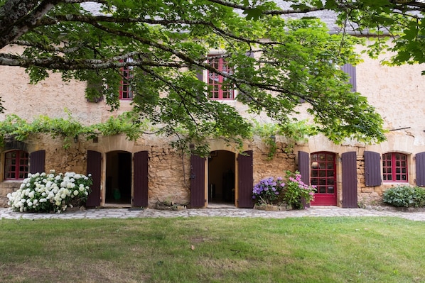 Un bâtiment en pierre, doté d’arcades et de volets rouges, entouré de verdure et bordé d’un chemin en pierre.