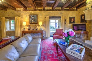 Une pièce aux murs de pierre, avec un canapé, un tapis rouge à motifs, un bureau avec des lampes, une chaise, un téléviseur et un vase rempli de fleurs.