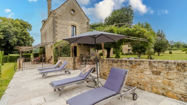 A stone building with a circular window, a pergola with lounge chairs, and a stone wall.